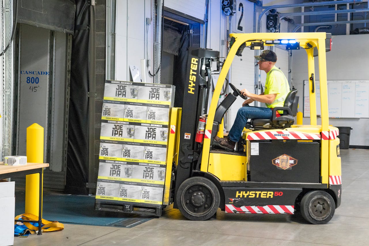 who-we-are A worker drives a Hyster forklift moving Joe IPA boxes in a warehouse.