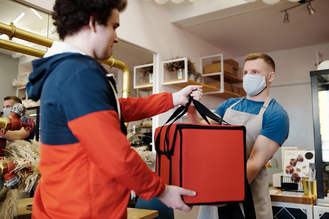 journey Two men exchanging a food delivery bag indoors, both wearing face masks.